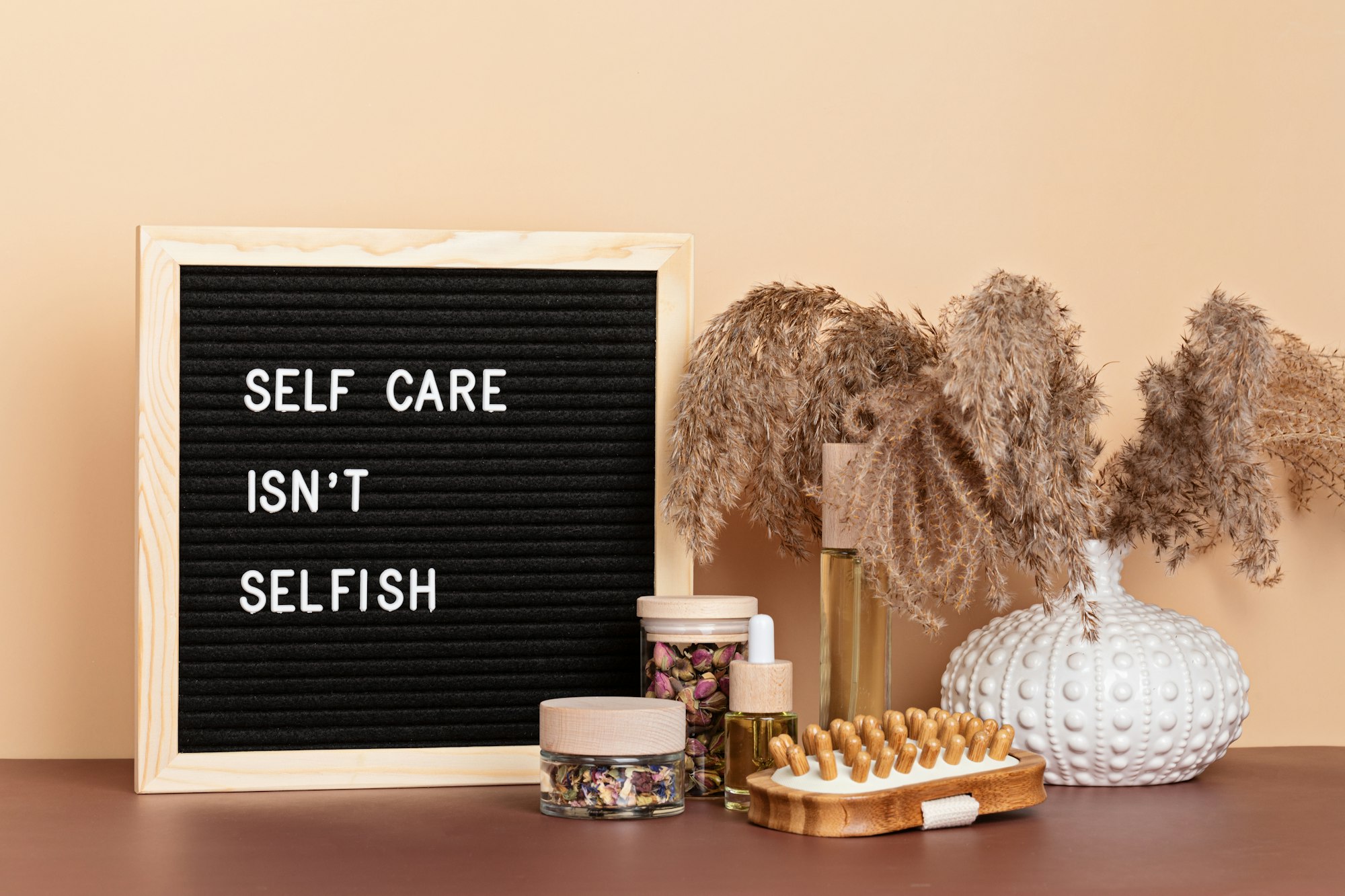 A letter board reading Self Care Isn't Selfish beside dried flowers, a face brush and jars of petals